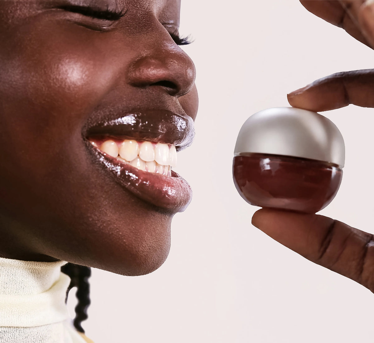 A close-up lifestyle shot of a woman smiling and licking gloss from a small, round jar held by another hand against a clean white background.